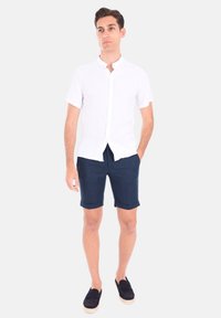 Maglietta bianca a maniche corte con colletto alla cinese, abbinata a pantaloni corti blu scuro. I pantaloni hanno i risvolti, e le scarpe nere completano l'outfit.