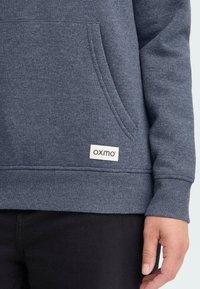 Pilkas džemperis su kengūros kišenę ir prigludusiomis rankovėmis. Apatinėje siūlės dalyje turi kvadratinę etiketę "oxmo".