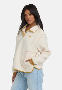 Frau in einem cremefarbenen Fleecepullover mit senfgelbem Besatz und hellblauen Jeans, die mit einer Hand in der Tasche vor einem weißen Hintergrund posiert.