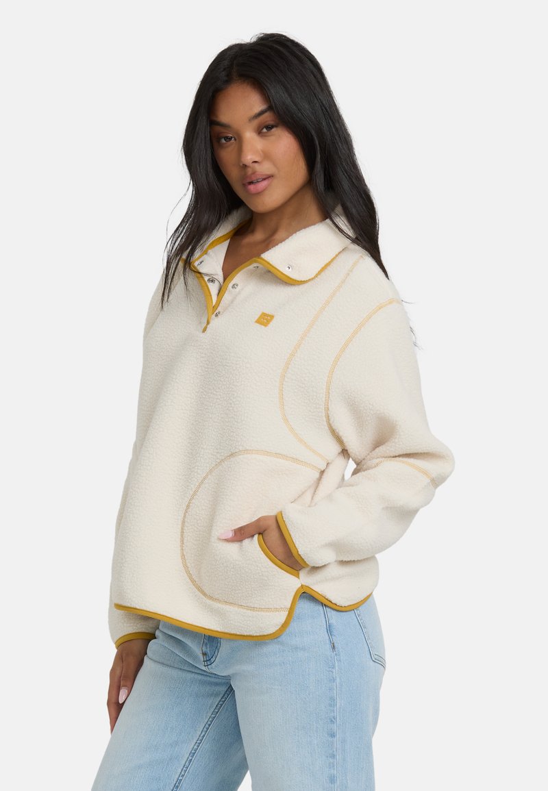 Frau in einem cremefarbenen Fleecepullover mit senfgelbem Besatz und hellblauen Jeans, die mit einer Hand in der Tasche vor einem weißen Hintergrund posiert.