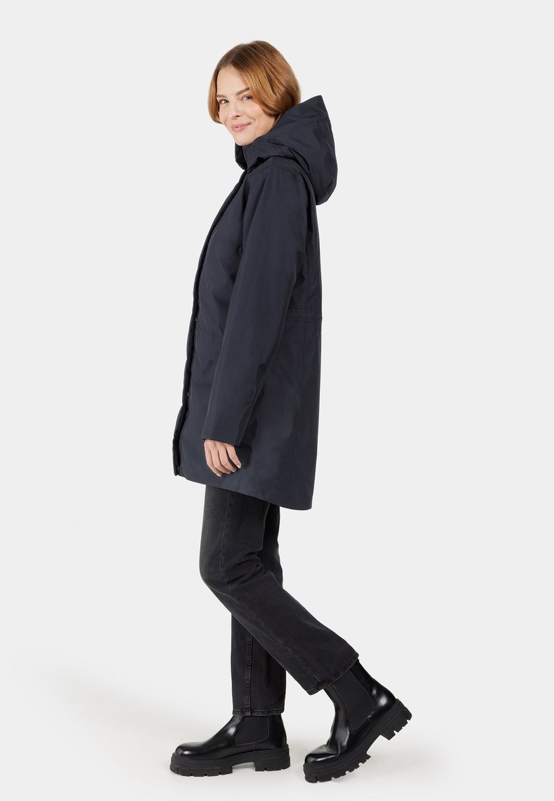 Langer, navyblauer, wasserdichter Mantel mit Kapuze, ausgestattet mit einer vorderen Leiste, Seitentaschen und geradem Schnitt, kombiniert mit schwarzen Jeans und klobigen Stiefeln.