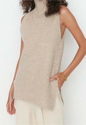 Kvinde iført ærmeløs beige strikket rullekravesweater med sidesnit, lyse bukser og guldkædearmbånd, hånd i lommen.