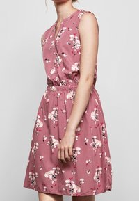 Ärmlöst klänning i dammig rosafarvet med vit och röd blommig tryck, knappstängning fram och elastisk midja, bärs av en stående person mot en enfärgad bakgrund.