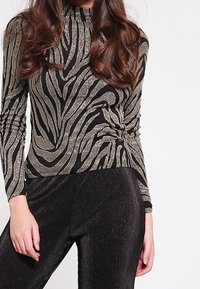 Czarna bluzka z długimi rękawami w metaliczne złote paski zebra, z wysokim dekoltem i bocznymi marszczeniami, zestawiona z czarnymi prążkowanymi spodniami.