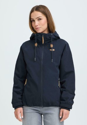 Valge taustaga naine, kellel on helepruunid juuksed ja kellel on seljas tumesinine kapuutsiga jakk, millel on ees lukk ja lukuga taskud.