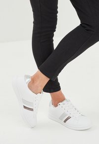 Zapatillas blancas con rayas grises y plateadas, con un acabado de cuero suave y un acento plateado en el talón. Combinadas con pantalones negros ajustados.