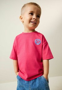 Lyserød t-shirt med korte ærmer og en blå smiley-logo på brystet, kombineret med blå denimshorts. Stoffet virker blødt og afslappet.