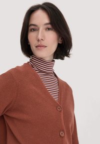 Vrouw met kort donker haar die een bruine gebuttonde cardigan draagt over een bruine en roze gestreepte coltrui tegen een effen lichte achtergrond.