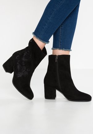 Schwarze Wildleder-Stiefeletten mit Blockabsätzen und seitlichen Reißverschlüssen, getragen zu blauen Skinny-Jeans mit ausgefranstem Saum. Ein Stiefel zeigt dezente Blumenstickerei.