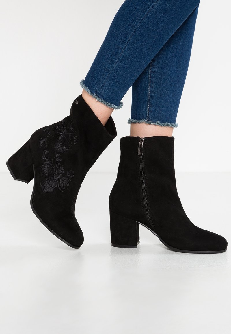 Bottines en daim noir avec talons carrés et fermetures éclair latérales, portées avec un jean skinny bleu à ourlets effilochés. Une des bottines présente une broderie florale subtile.