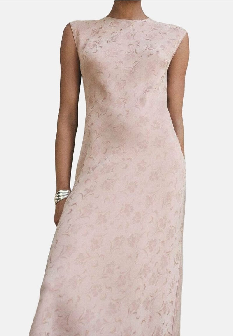 Femme portant une robe sans manches rose pâle avec un motif floral subtil et un bracelet argenté au poignet gauche sur un fond uni.