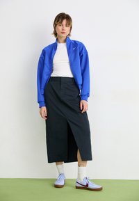 Blå sportsjakke med lynlås, hvid top, sort lang nederdel med sideslids og lyseblå sneakers med brun sål og røde accenter.