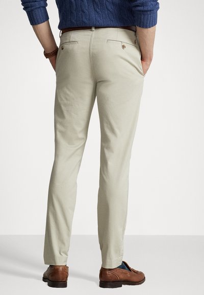 Beige bomullschinos med slim fit, med to baklommer med knappelukking. Kombinert med brune lær sko, sett fra baksiden.