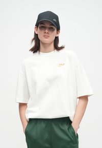 Jauns cilvēks ar taisniem brūniem matiem, valkā melnu cepuri, baltu lielu T-kreklu ar tekstu "Vienkārši dari to" un tumši zaļas bikses, rokas kabatās.