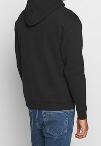 Uomo con un hoodie nero e jeans blu, visto da dietro su uno sfondo grigio chiaro uniforme.
