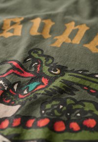 La tela verde presenta un diseño de dragón con acentos rojos y letras doradas, mostrando escamas detalladas y un ojo prominente.