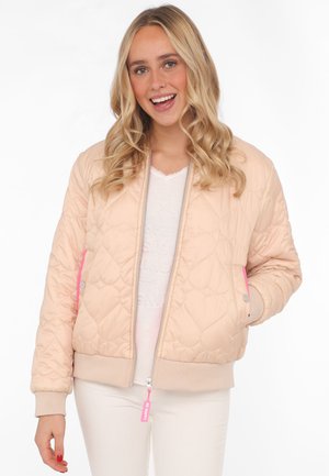 Junge Frau mit blondem lockigem Haar, die eine hellrosa gesteppte Jacke mit Herzmustern und weiße Hose trägt, lächelt vor einem weißen Hintergrund.