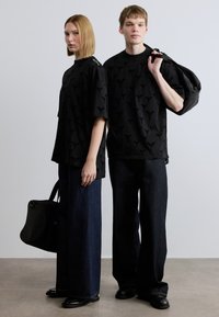 Μαύρα κοντομάνικα πουκάμισα με υφή, σε συνδυασμό με φαρδιά σκούρα τζιν παντελόνια. Ο άνδρας μοντέλο φοράει μια μαύρη τσάντα.