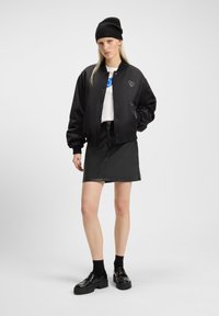 Black bomber jacket with heart detail, white t-shirt, black leather mini skirt, and glossy platform shoes. Model wearing a black beanie.