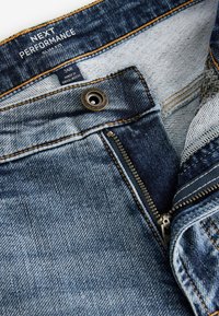 Modré rifle z denimu v slim fit strihu, s žltým prešitím, kovovým zipsom a pásovou nášivkou s nápisom "NEXT PERFORMANCE."