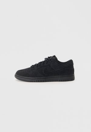 Schwarzer Low-Top-Sneaker aus Wildleder mit Schnürsenkeln, Gummisohle, perforiertem Zehenbereich und dezentem Logo an der Seite, in Seitenansicht vor weißem Hintergrund gezeigt.