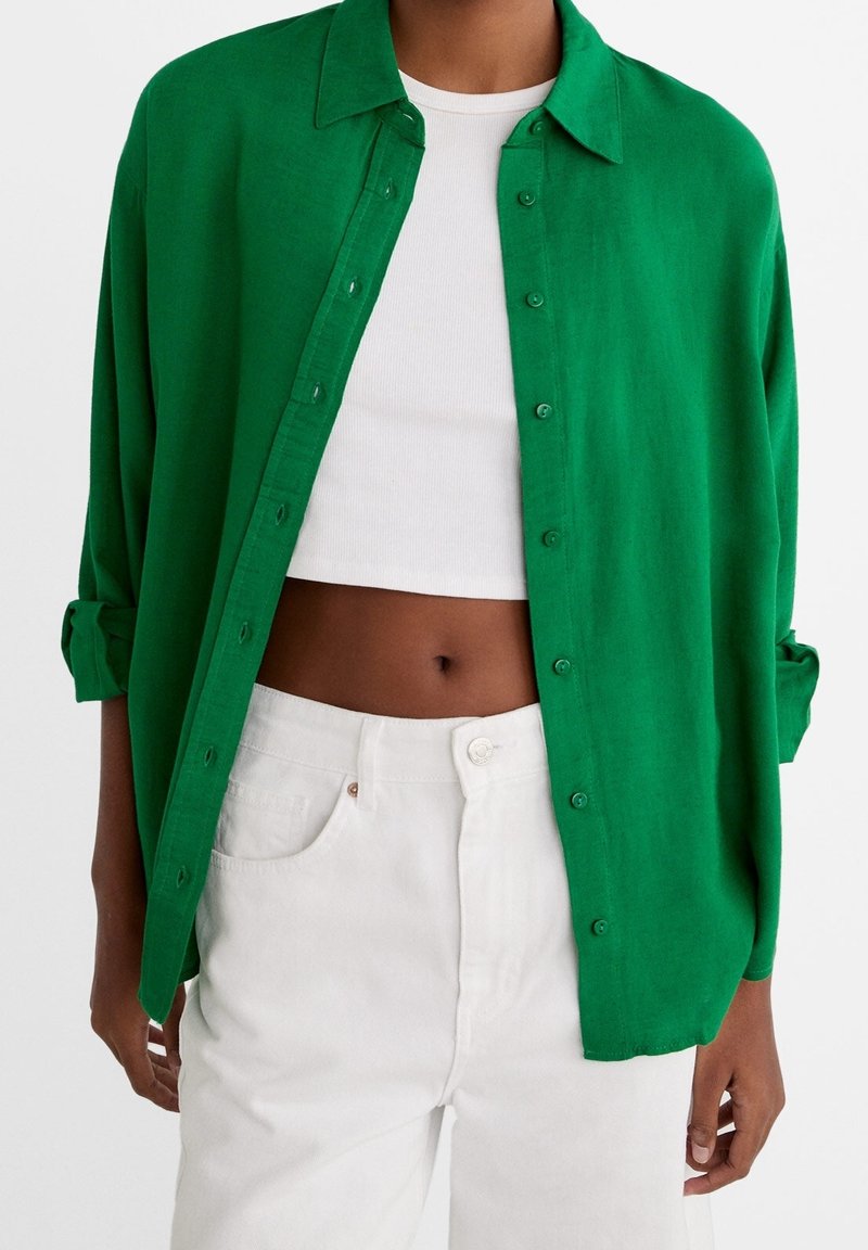 Person wearing a bright green button-up shirt over a white crop top, paired with high-waisted white pants against a plain background.