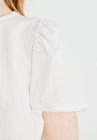 Gros plan d'une personne portant un haut blanc avec des manches courtes bouffantes et un galon en dentelle festonnée sur un fond uni.
