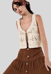 Gilet au crochet de couleur beige clair avec des motifs floraux et une fermeture à nouer, associé à une jupe marron à boutons avec un ourlet froncé.
