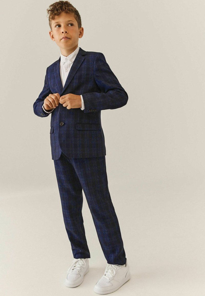 Marineblauwe geruite pak met een enkelknopsjasje en notch-lapels, gecombineerd met een witte dress shirt en witte sneakers. Soepele textuur.