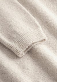 Weicher, cremefarbener Strickstoff, der die Manschette eines langärmligen Pullovers mit gerippter Struktur und sichtbaren feinen Wollfasern zeigt.