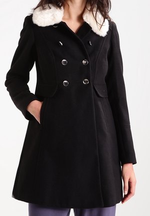 Femme portant un manteau noir croisé à boutons argentés et un col en fausse fourrure blanche, main dans la poche, sur fond blanc.