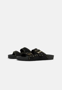 Infradito in suede nero con due fibbie regolabili, dettagli in borchie dorate, suole testurizzate e design piatto.