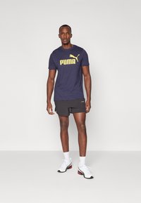 T-shirt de sport bleu marine avec le logo "PUMA" jaune, shorts noirs, baskets blanches avec des accents rouges et chaussettes blanches.