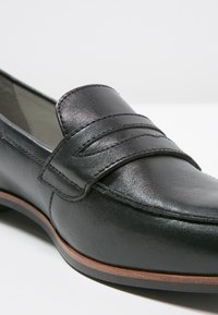 Mocassin en cuir noir avec un bout arrondi, une bride décorative sur le dessus et une semelle contrastante marron et noire.