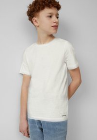 Teenager mit lockigem rotem Haar, der ein schlichtes weißes Kurzarm-T-Shirt und helle blaue Jeans trägt, steht an einer grauen Wand.