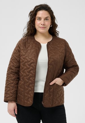Femme portant une veste matelassée marron zippée sur un haut blanc et un pantalon noir, avec une main dans la poche de la veste sur un fond uni.