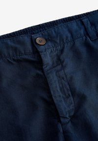 Pantaloni blu navy con vita elasticizzata, chiusura con bottone marrone e texture liscia. Presentano tasche laterali e cuciture curate.