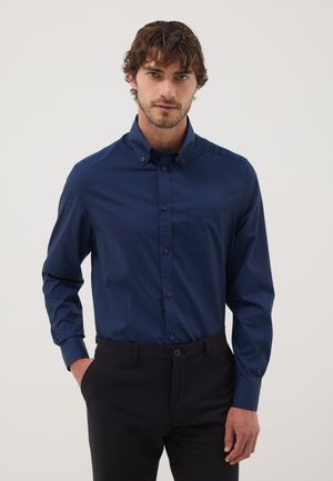 Uomo con capelli ricci che indossa una camicia blu scuro con bottoni e pantaloni neri, in piedi con una mano in tasca davanti a uno sfondo semplice.