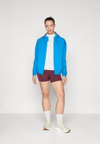 Blue lightweight jacket, white top, maroon shorts, and beige sneakers. Textured fabric with a zippered pocket and hood detail.