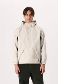 Beige pullover jacket with a hood, featuring a front pocket, navy side panels, and snap buttons. Made of smooth, lightweight fabric.