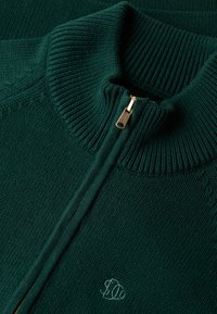 Donkergroene gebreide zip-up trui met een geribde kraag en manchetten, voorzien van een gouden rits en een geborduurd logo op de voorkant.