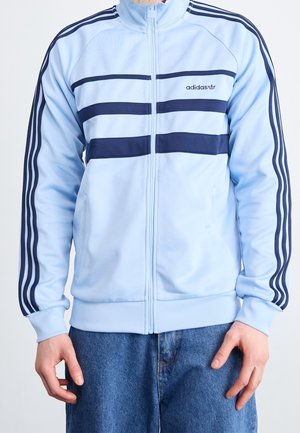 Person iført en lyseblå Adidas lynlåsjakke med mørkeblå striber på ærmerne og brystet, kombineret med blå denimjeans.