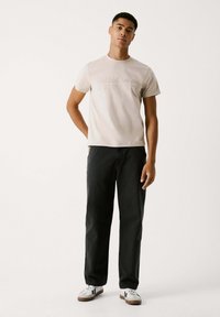 Beige bomulds T-shirt med diskret branding, parret med mørke denimbukser og hvide sneakers med sorte accenter. Casual design, afslappet pasform.