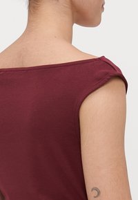 Persona que lleva una blusa sin mangas y con los hombros al descubierto de color burdeos, mostrando la parte superior de la espalda, el hombro y un pequeño tatuaje de luna creciente en el brazo.