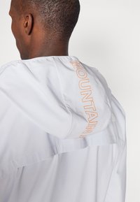 Balta lengva striukė su kapuce, oranžine "MOUNTAIN" užrašu ant nugaros ir subtiliu logotipo detalėmis viršutinėje dešinėje petyje.