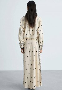 Ensemble beige assorti comprenant une longue jupe et une chemise surdimensionnée, ornés de petits motifs de palmiers noirs et d'accents décoratifs sur les côtés.