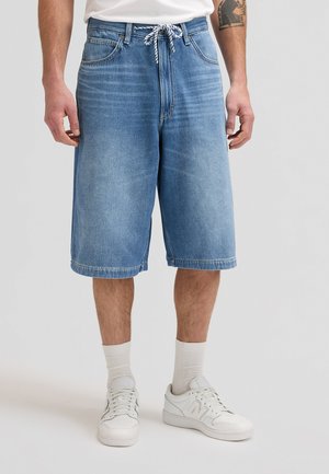 Homme portant un short en jean bleu clair avec taille à cordon, chaussettes blanches et baskets blanches, debout devant un fond uni.
