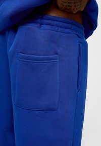 Blauwe joggingbroek van zachte stof heeft een zichtbare zijzak en een elastische tailleband. De textuur lijkt glad en consistent.