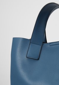 Modrá kožená kabelka přes rameno s texturovaným povrchem. Má strukturovaný tvar a široký držák, s jemnými švy.