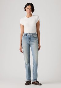 Άσπρο ριμπ κοντομάνικο τοπ συνδυασμένο με ανοιχτό μπλε ψηλόμεσο τζιν. Το τζιν έχει ίσια γραμμή και ελάχιστη ξεθώριαση· το μοντέλο φοράει σκουρόχρωμα παπούτσια.